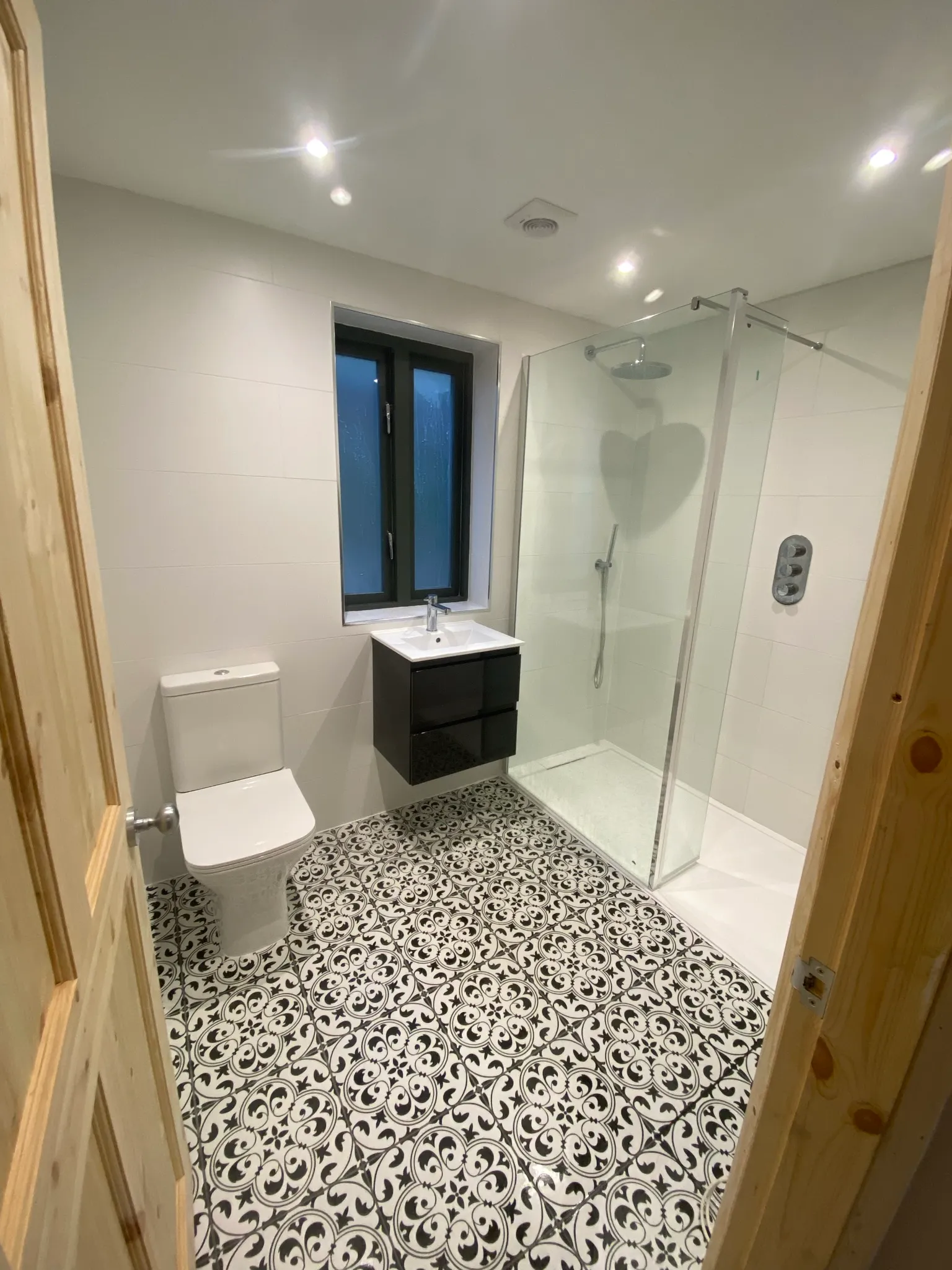 Finished bathroom with white-tiled walls, a wall-hung black gloss vanity, a glass-screened walk-in shower and a black-and-white patterned floor.