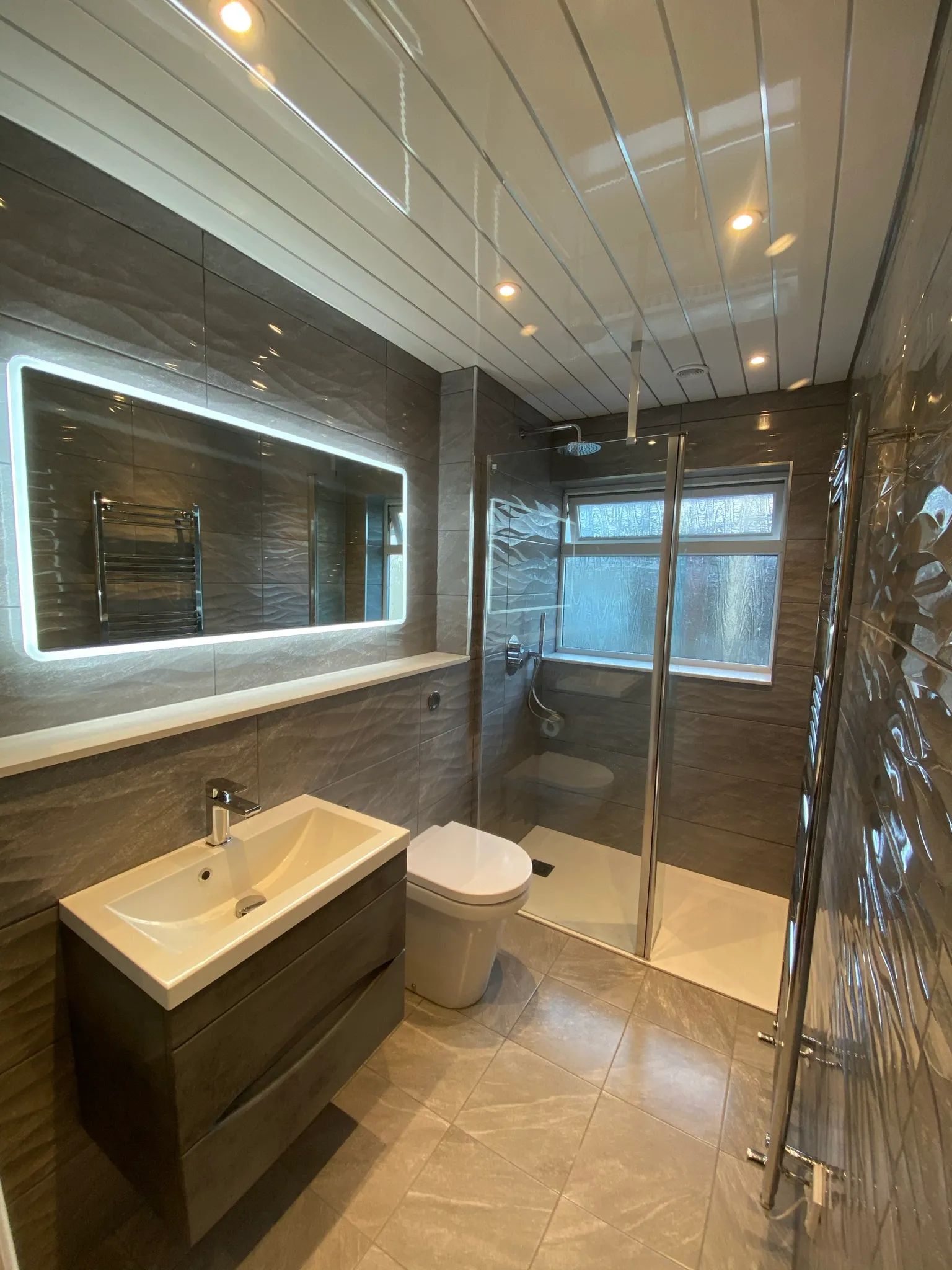 Finished bathroom with grey wave-textured tiling, a large illuminated LED mirror, a wall-hung vanity and a walk-in glass shower.