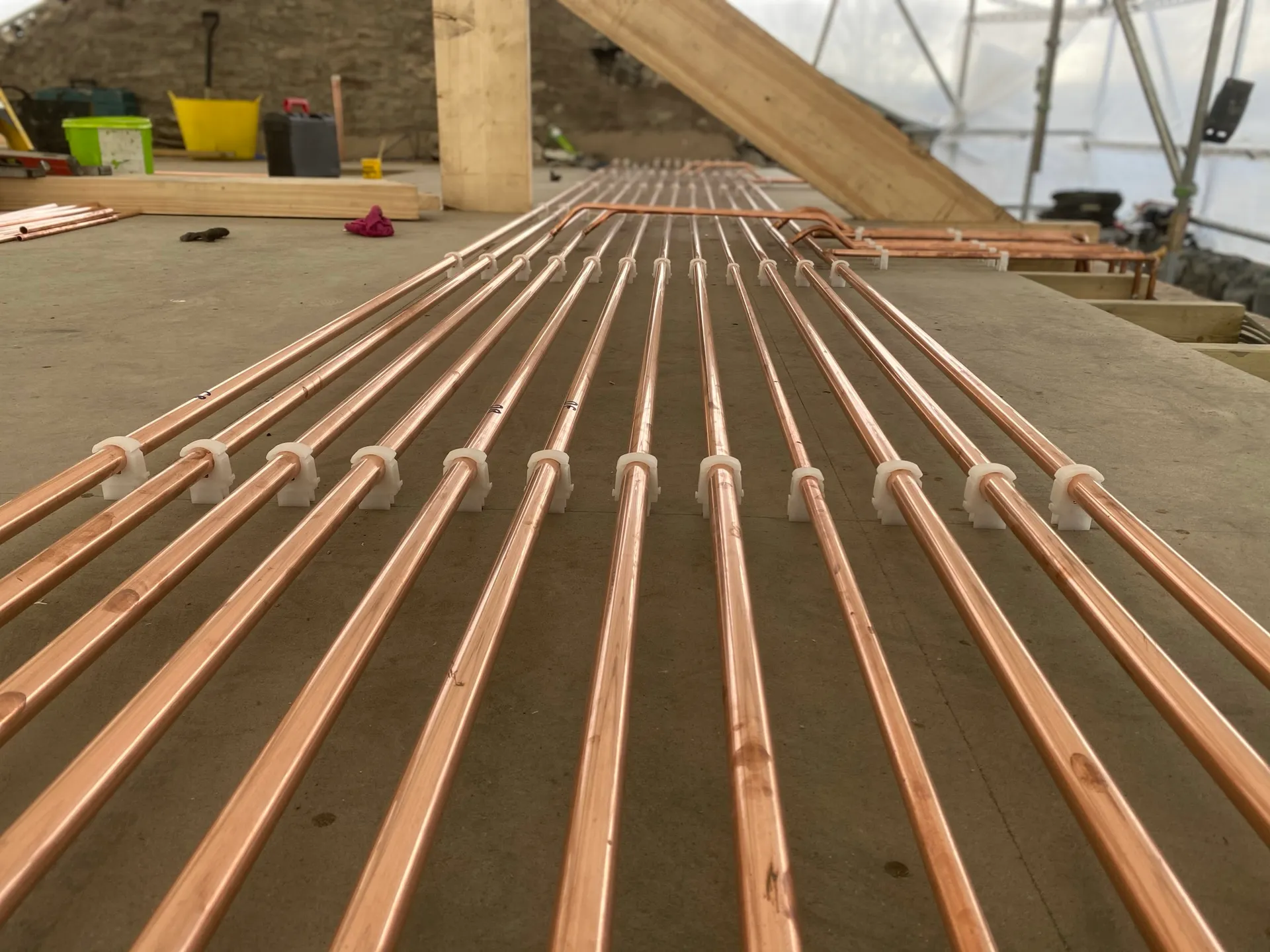 Low-angle view along many parallel new copper pipes held in white clips, receding across a concrete floor.