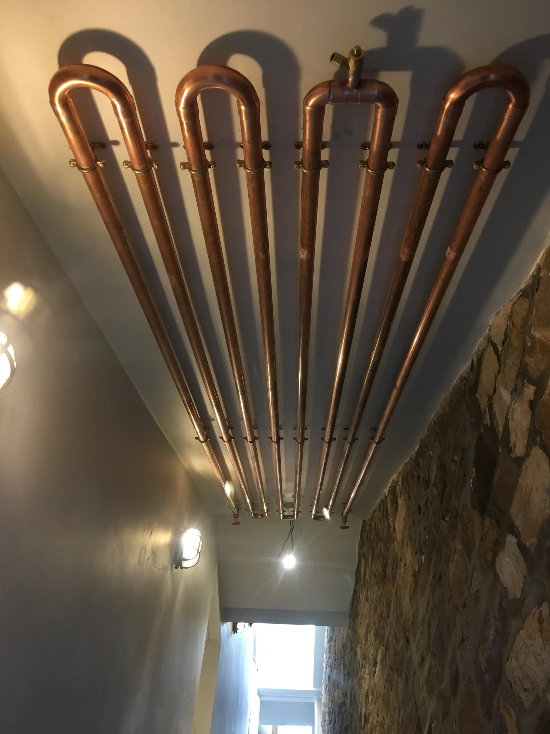 Dramatic low-angle view of a polished copper serpentine pipe feature against a white wall beneath an exposed stone ceiling.