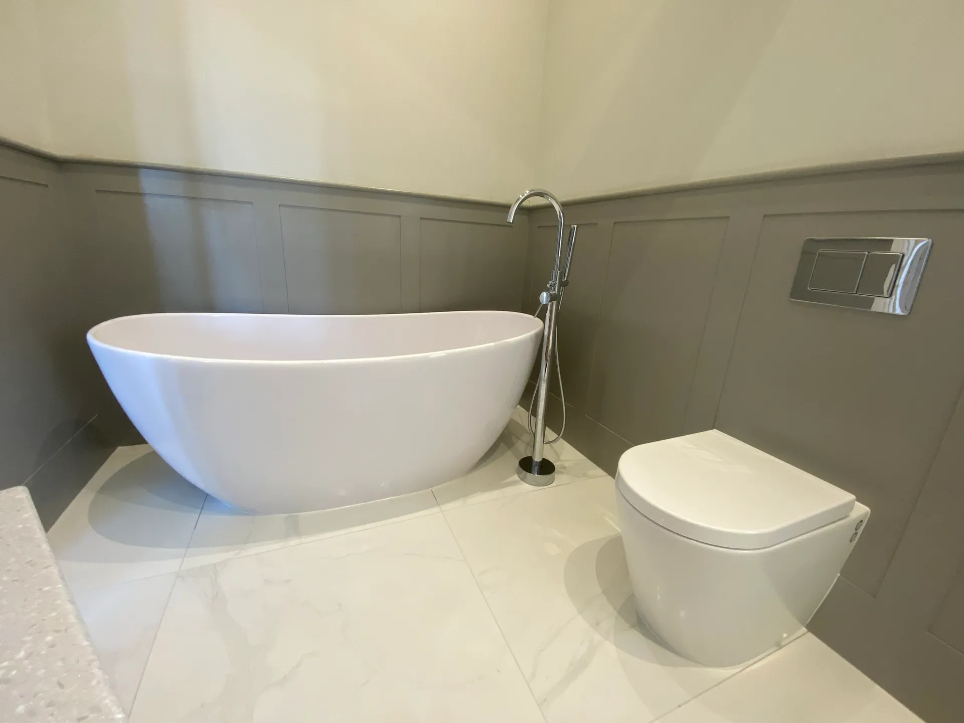 Freestanding white slipper bath with a floor-standing chrome bath and shower mixer beside a back-to-wall toilet, against grey shaker wall panelling.