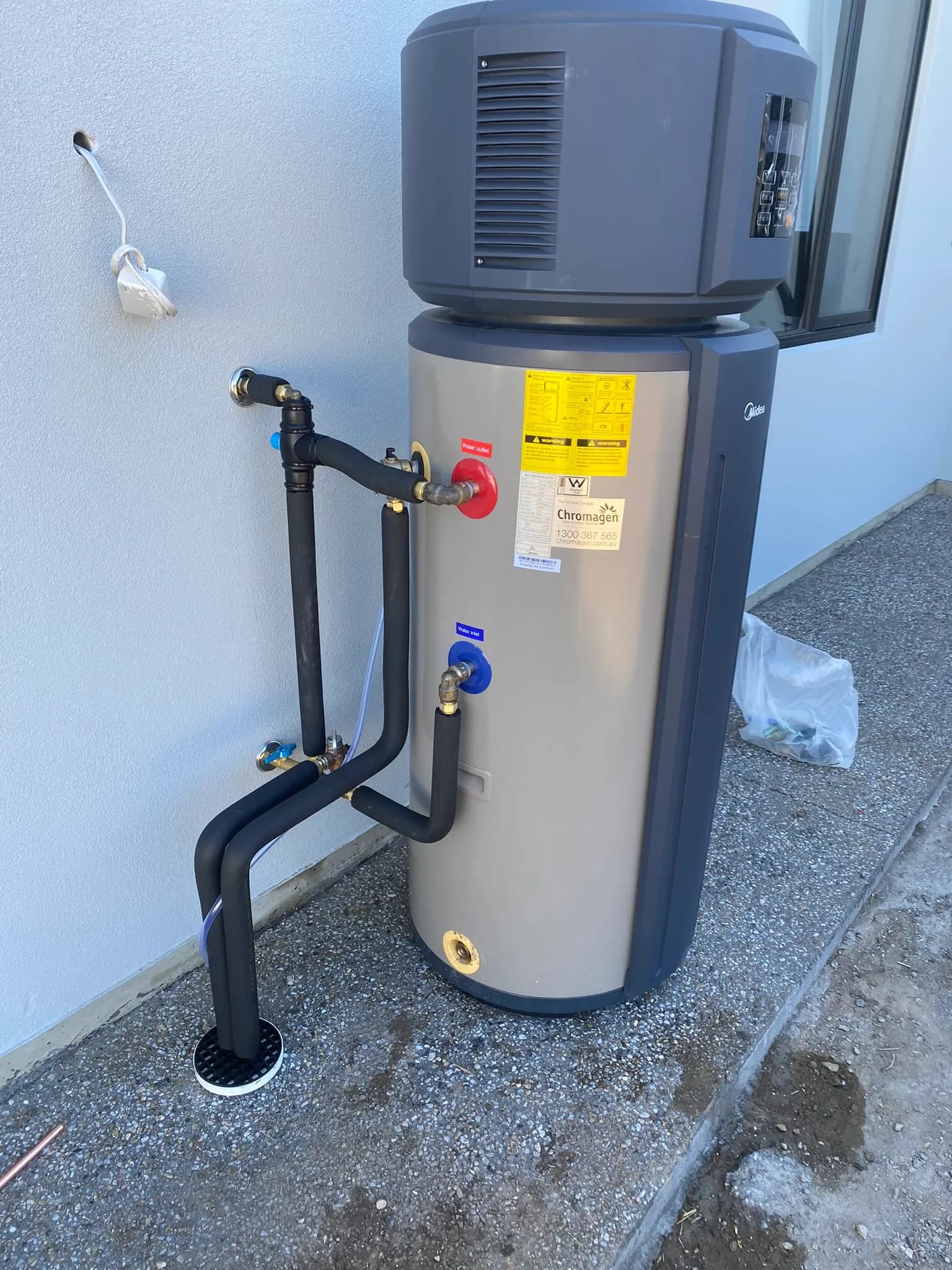 Outdoor heat-pump hot water system with a grey storage cylinder and heat-pump unit connected with lagged pipework.