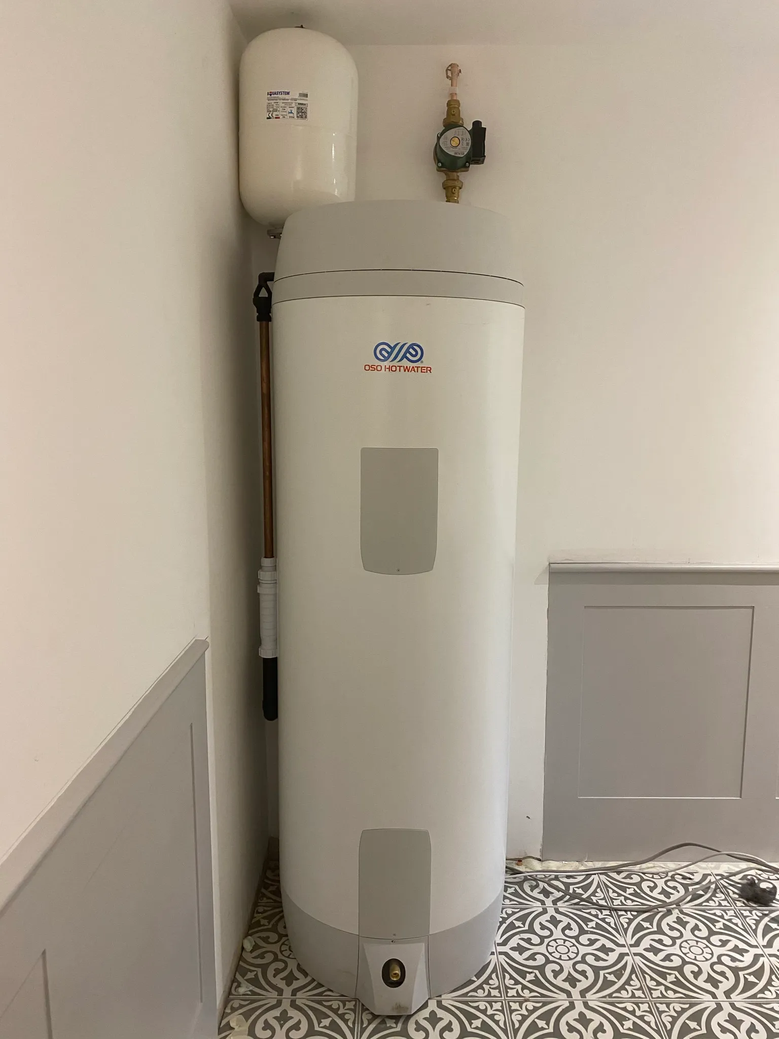 White unvented hot water cylinder installed with an expansion vessel and copper pipework in a bright corner.