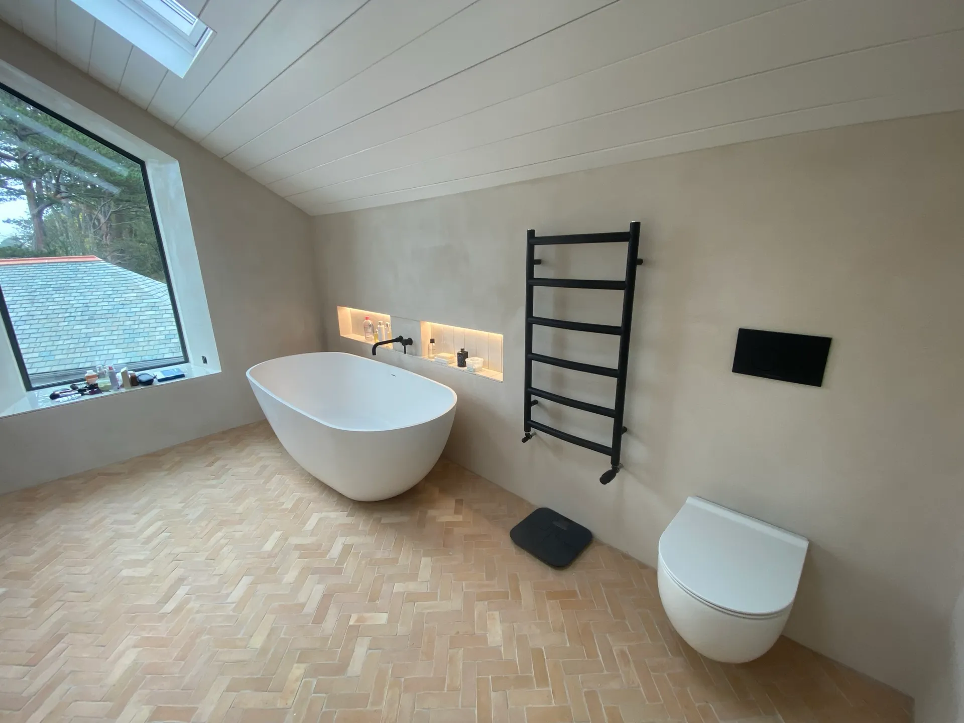 Loft bathroom with a freestanding bath beneath an angled window, microcement walls, a black ladder towel radiator and a wall-hung toilet.